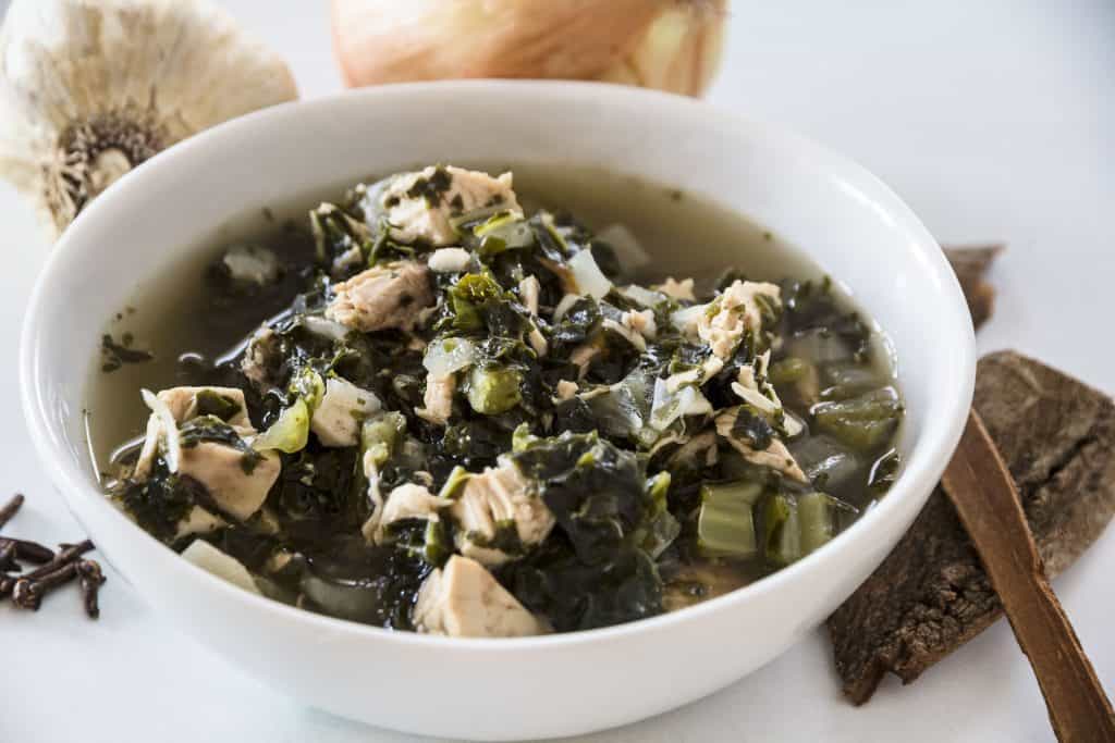 sideways shot of low carb chicken kale soup in a white bowl, with onions, garlic, cinnamon and cloves in the background