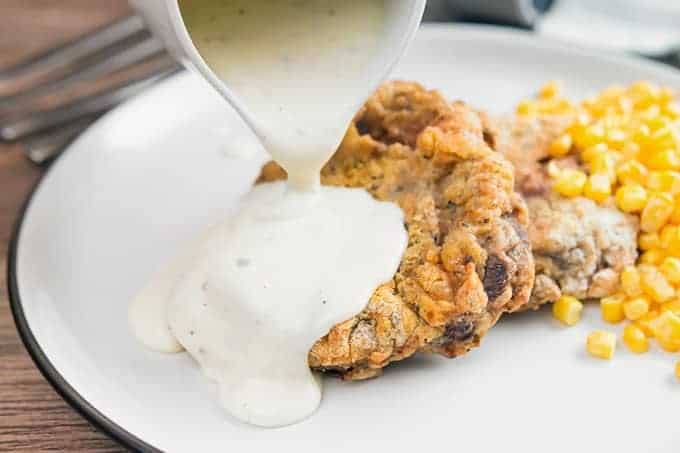 Chicken Fried Steak with country gravy poured on top with corn on the side on a white place. 