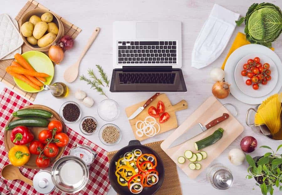 Home kitchen table top view with laptop, food ingredients, raw vegetables, kitchenware and utensils, top view