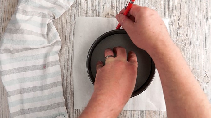 Two hands tracing a round pot with a pencil on parchment paper.
