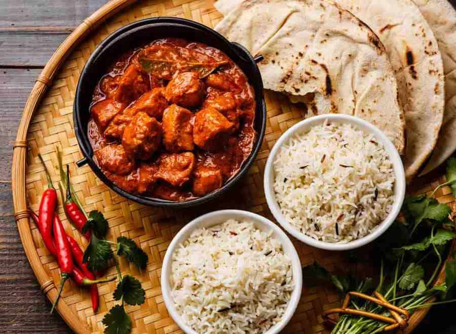 A bowl of Chicken Tikka Masala with two bowls of rice and Naan bread on a table.