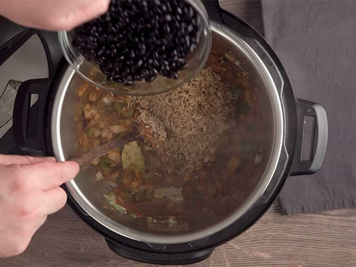 pouring black beans into an instant pot filled with uncooked brown rice and vegetables and spices
