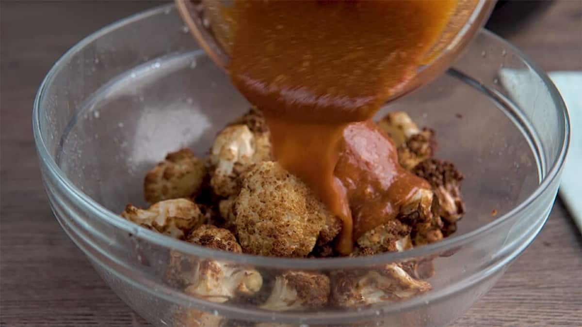pouring buffalo sauce over cooked cauliflower florets in a clear mixing bowl
