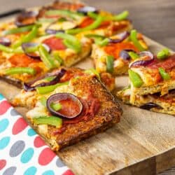 Slices of low carb pizza topped with onions, salami, peppers, and cheese on a cutting board