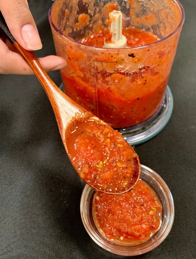 Overhead image of keto pizza sauce in a glass bowl being served with a wooden spoon held by a manicured hand. 