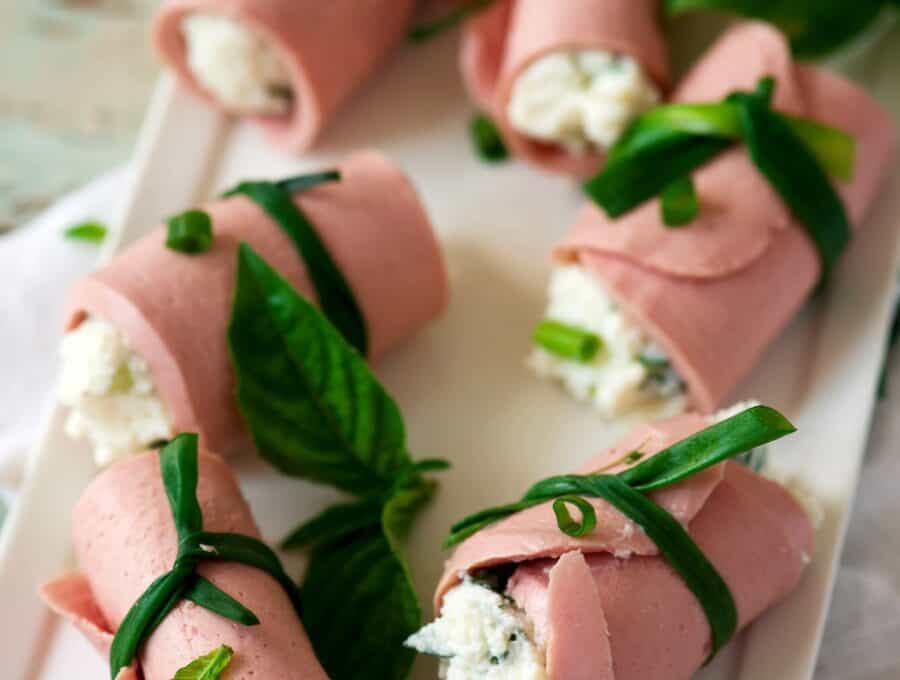 Rolls of deli meat wrapped around soft cheese and tied together with green onions on a white platter.