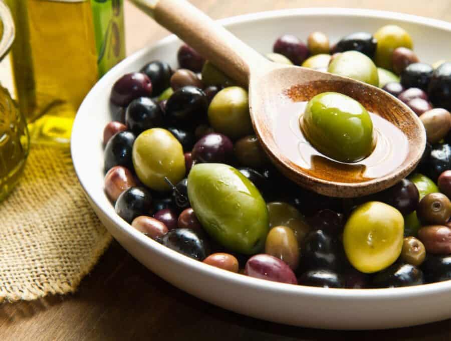 Image of green and black olives in a white bowl. A single olive in oil is held over the bowl in a wooden spoon.