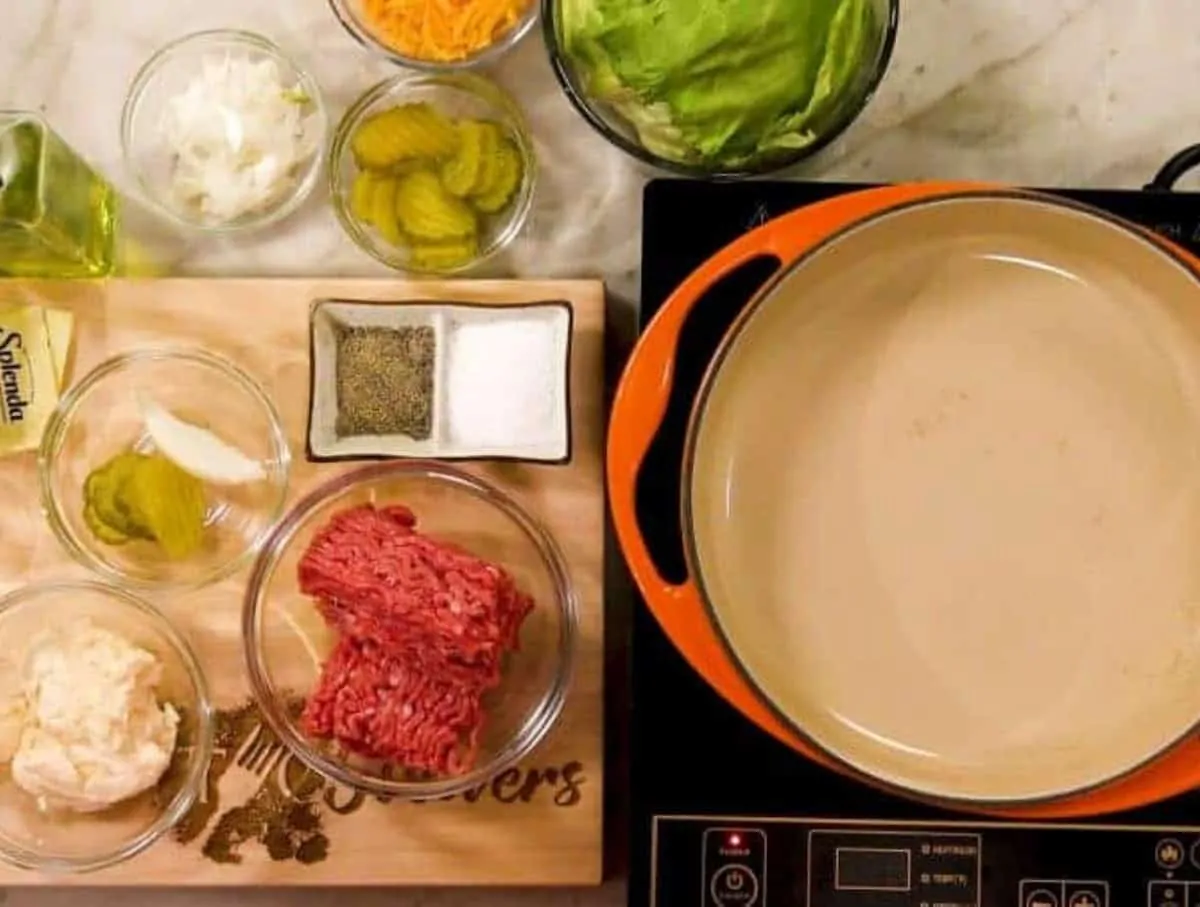 An orange ceramic pan on an induction burner with ground beef, mayo, pickles and onions, and salt in pepper in separate bowls to the side.