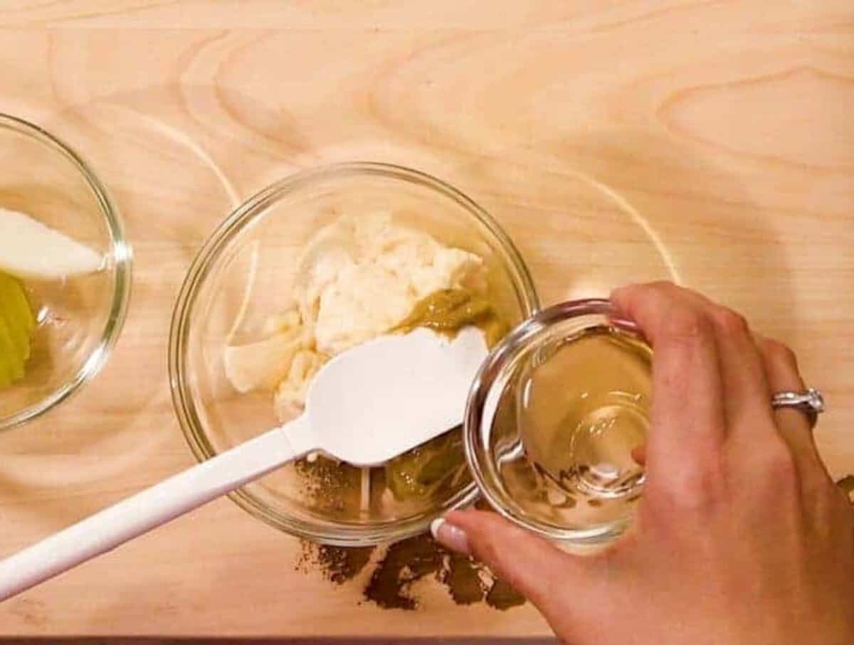 Hands pouring vinegar into a dish with mayo and mustard with a white spatula.