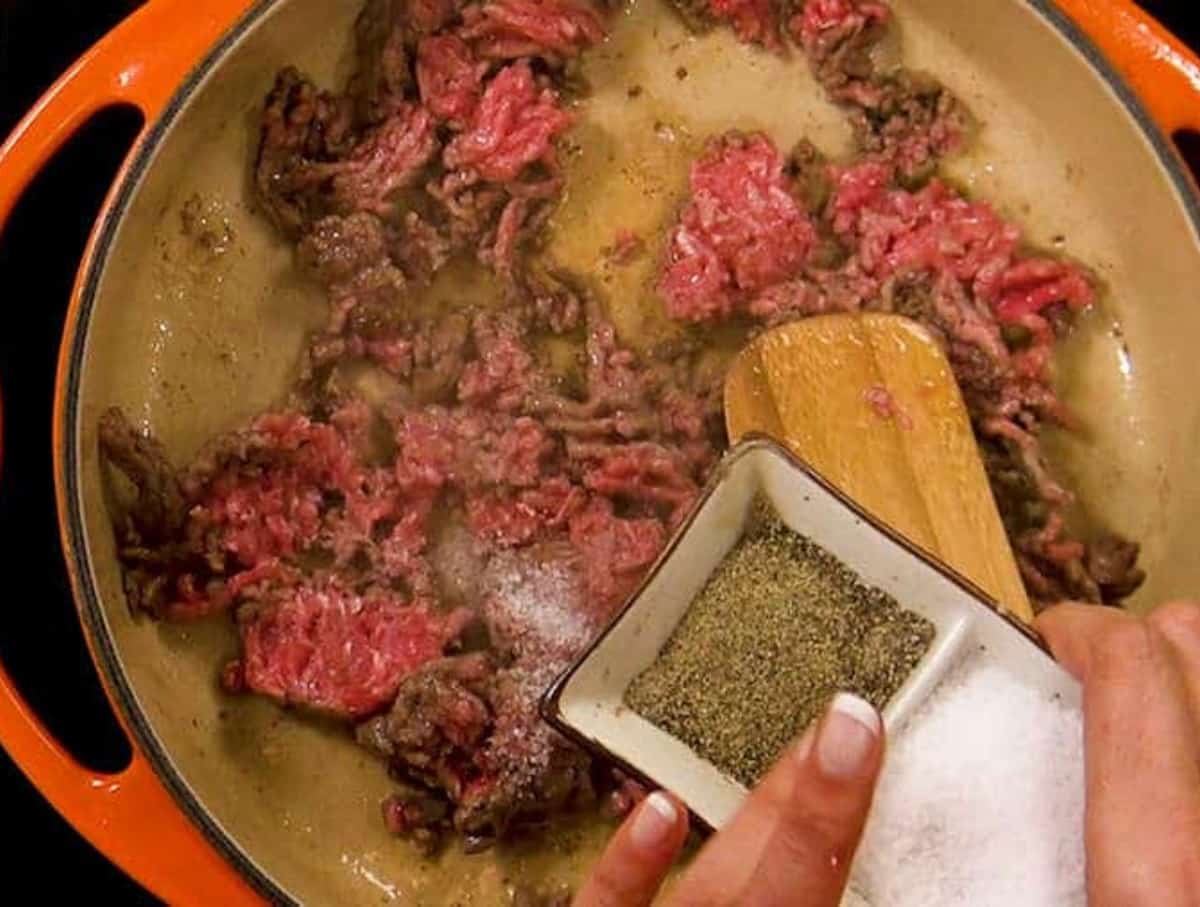 Fingers pushing salt and pepper into a ceramic pan on to cooked ground beef.