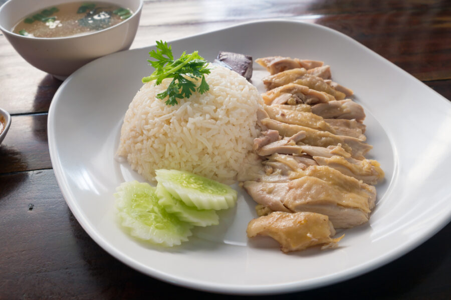 Hainanese Chicken Rice dish served on a white plate with a bowl of soup in the background.