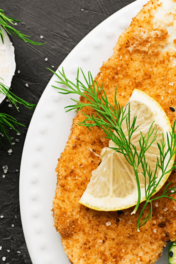 Close up image of chicken milanese on a white plate with a dill and lime garnish