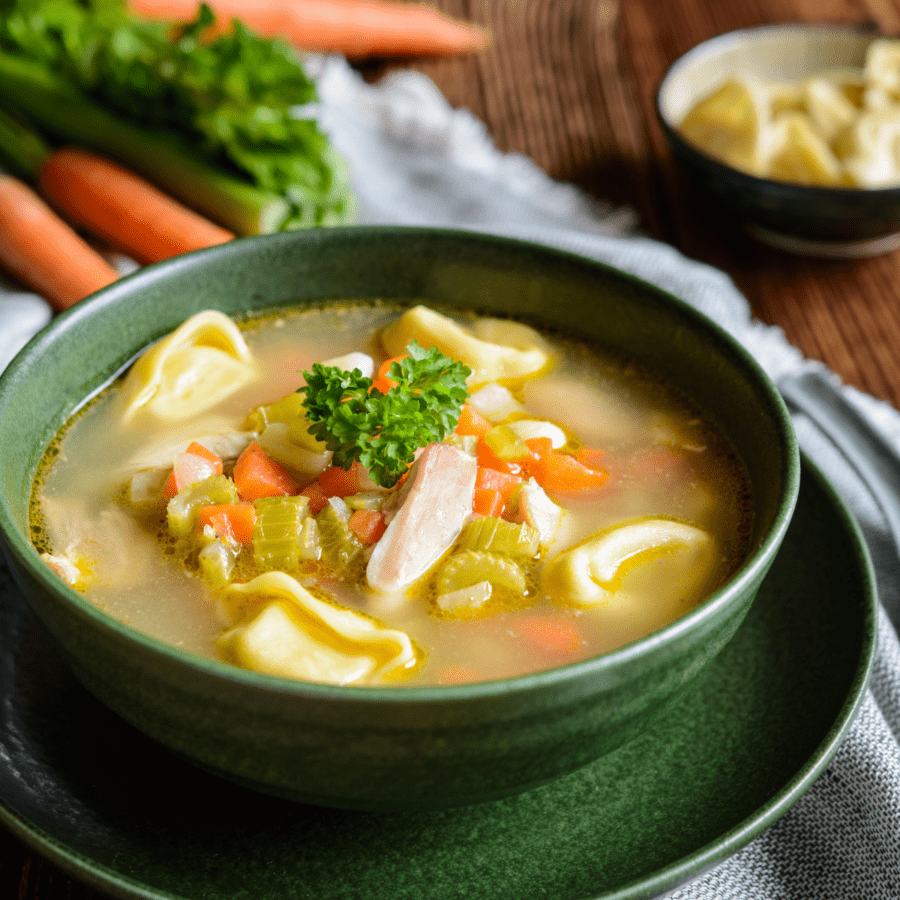 Chicken Tortellini Soup in a bowl sitting on a saucer with a blue napkin and carrots in the background