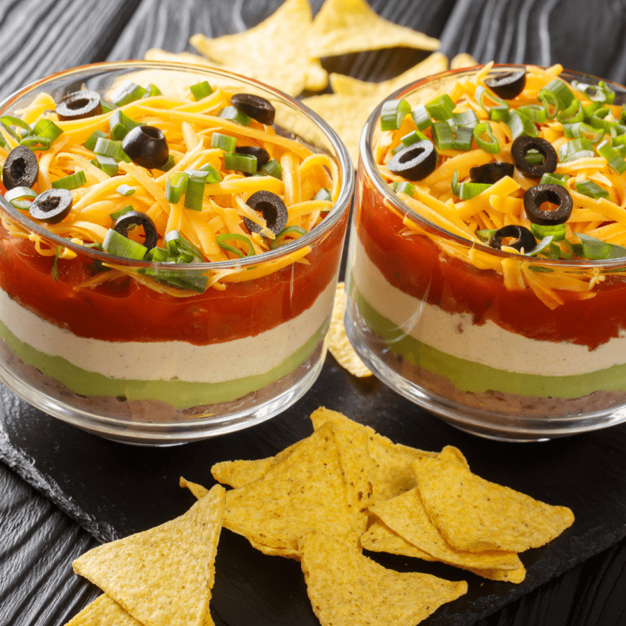 Two individual servings of taco dip on a black surface surrounded by tortilla chips