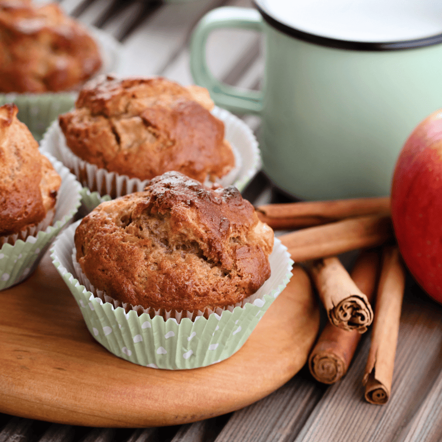 Apple cinnamon muffins on a wooden cutting board next to cinnamon sticks, apples, and a turquoise coffee mug