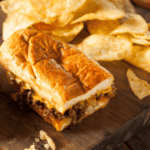 Close Up of a Chopped Cheese Sandwich on a wooden cutting board with a side of kettle chips