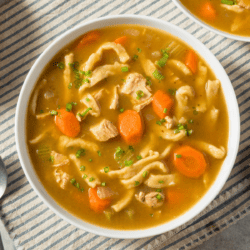 Close up image of a white bowl of chicken noodle soup on a striped backdrop