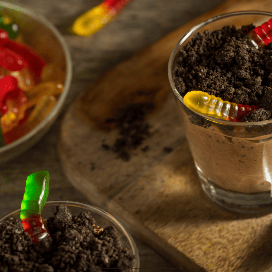 Close up image of two dirt cake cups on a wooden cutting board next to a bowl of gummy worms