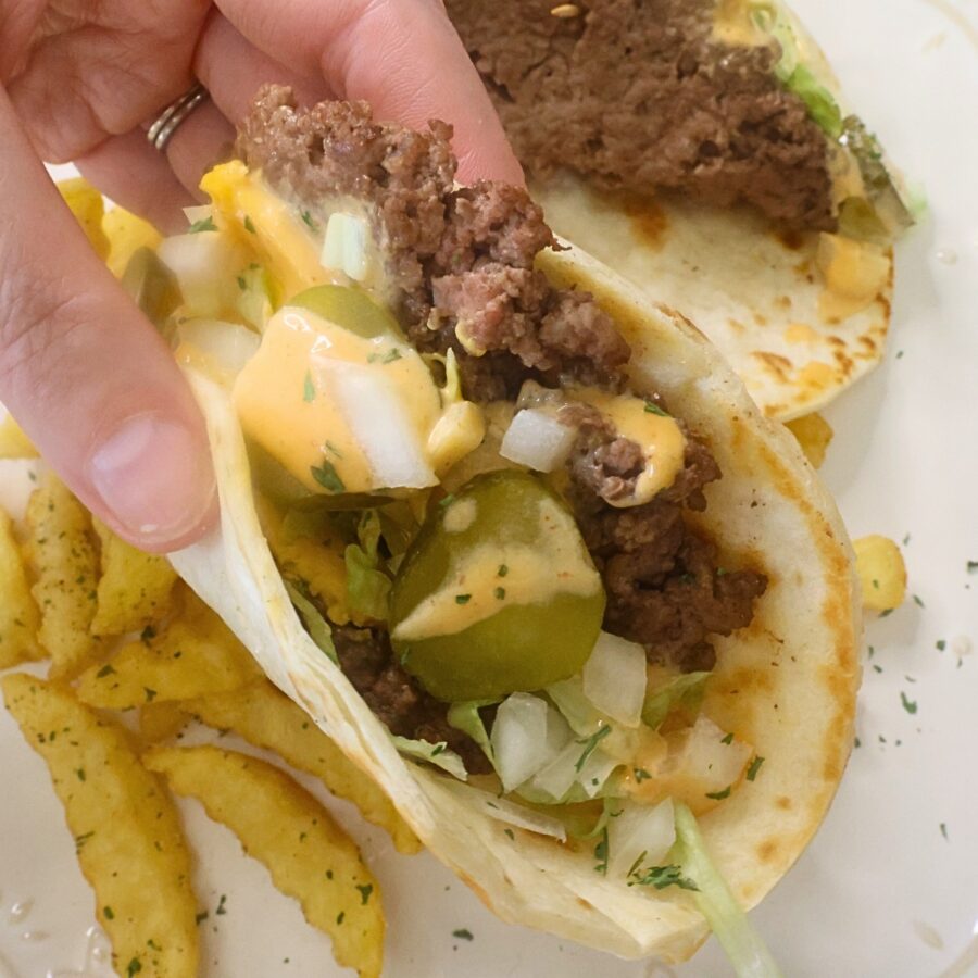 A hand holding a tortilla burger with fries on a plate in the background