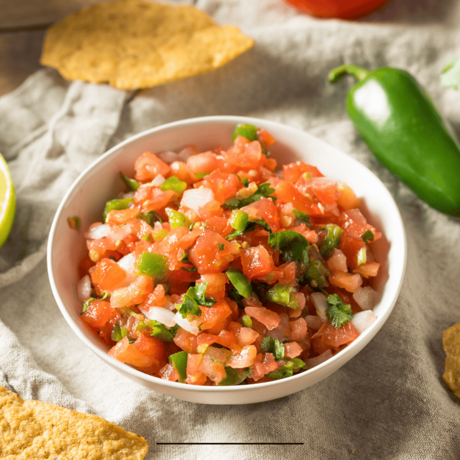 Pico de gallo in a white bowl surrounded by peppers and tortilla chips