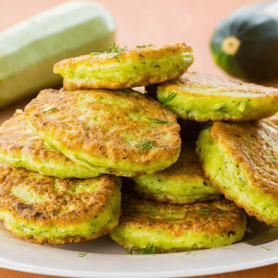 A large stack of zucchini pancakes on a white plate with whole zucchini in the background