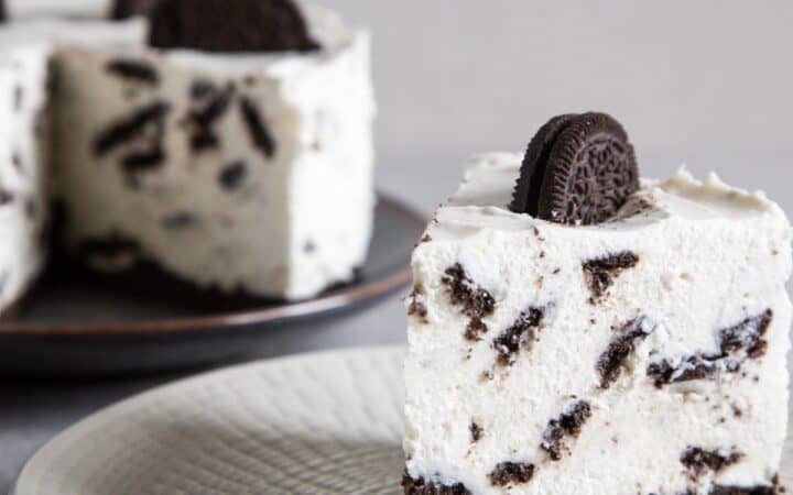 A slice of oreo cheesecake on a white plate with the remaining cheesecake in the background