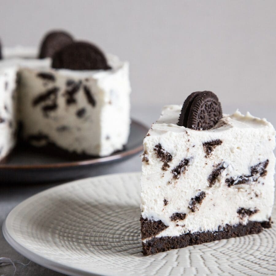 A slice of oreo cheesecake on a white plate with the remaining cheesecake in the background