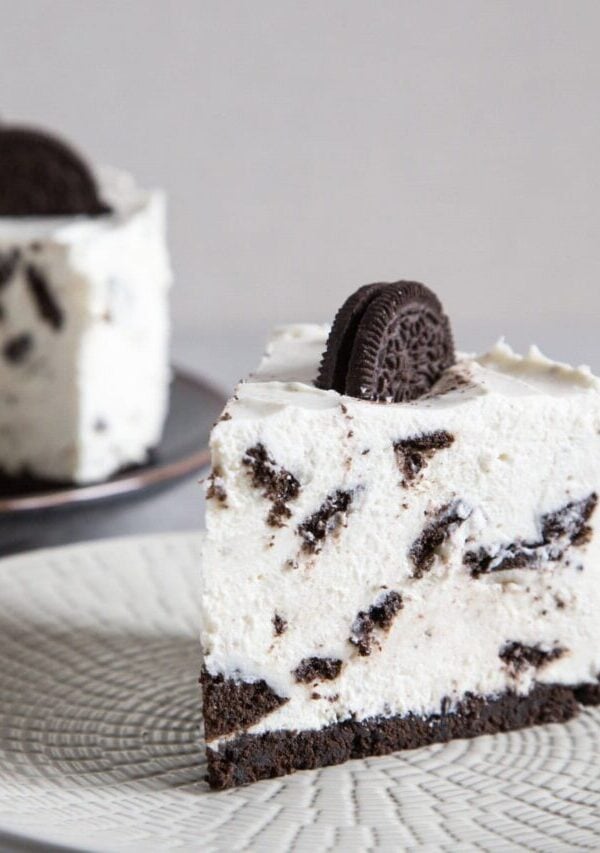 A slice of oreo cheesecake on a white plate with the remaining cheesecake in the background