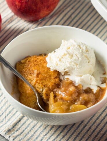 A white bowl full of Apple Cobbler and a scoop of vanilla ice cream