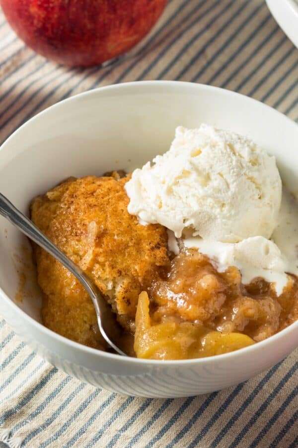 A white bowl full of Apple Cobbler and a scoop of vanilla ice cream