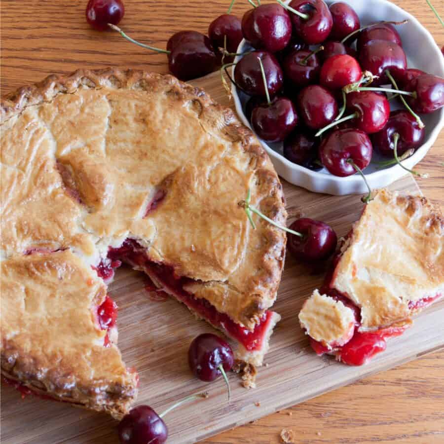 Overhead image of a cherry pie with a slice taken out of it on a wooden cutting board