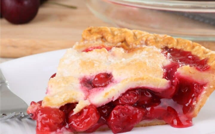 A slice of Cherry Pie on a white plate with a pie in the background