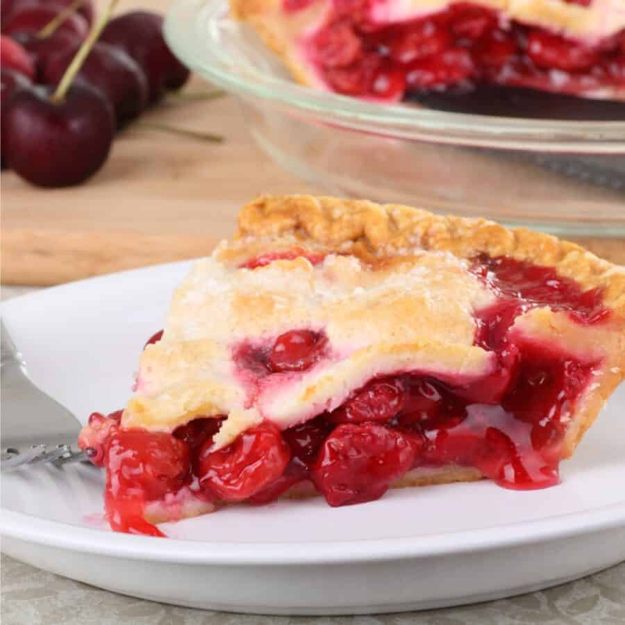A slice of Cherry Pie on a white plate with a pie in the background