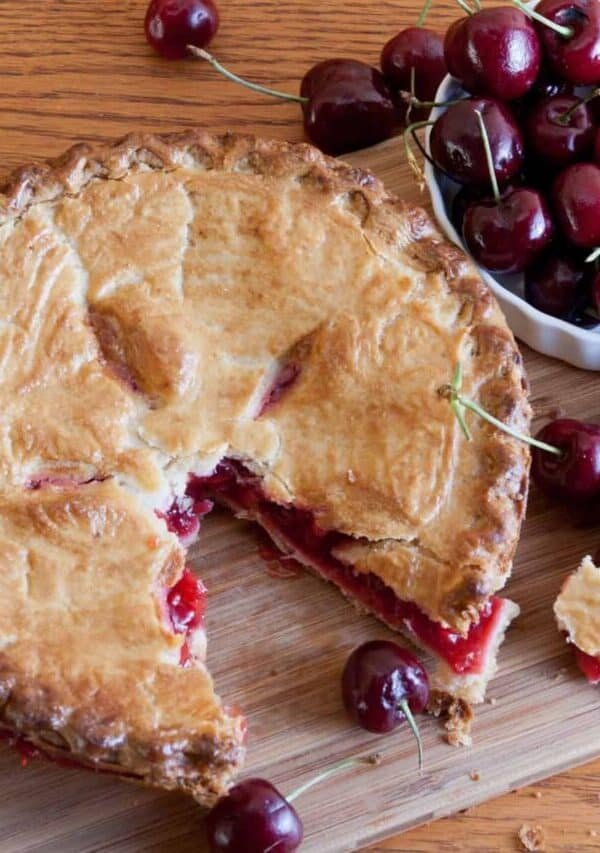 Overhead image of a cherry pie with a slice taken out of it on a wooden cutting board