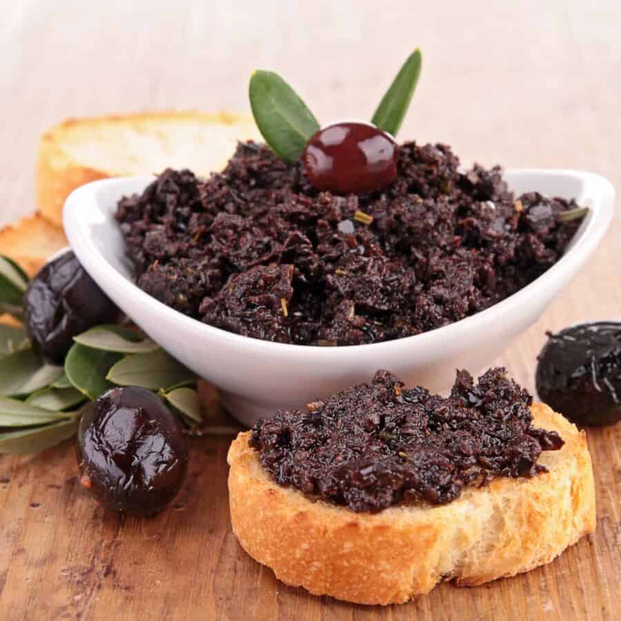 Olive Tapenade in a white bowl and spread on a piece of bread