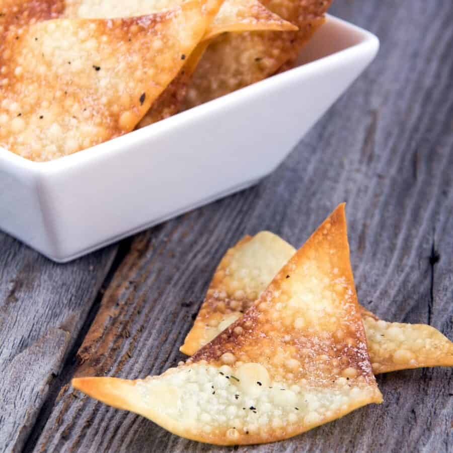 Two Baked Wonton Chips on a rustic surface next to a bowl full of them