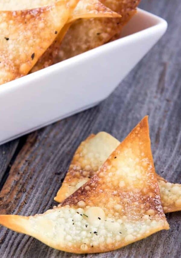 Two Baked Wonton Chips on a rustic surface next to a bowl full of them