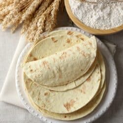 Overhead image of Flour Tortillas stacked on a white plate next to a bowl of flour