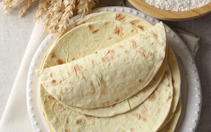 Overhead image of Flour Tortillas stacked on a white plate next to a bowl of flour