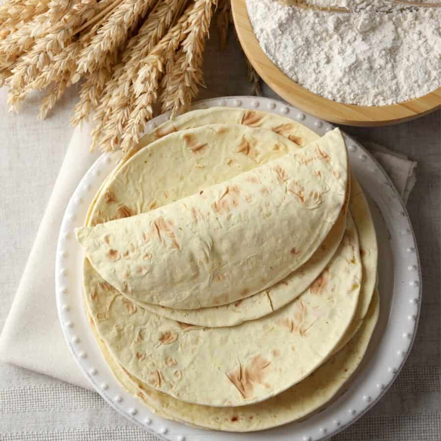 Overhead image of Flour Tortillas stacked on a white plate next to a bowl of flour