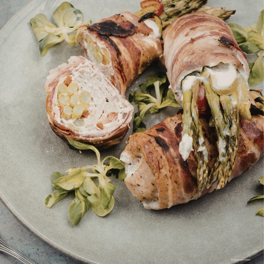 Close up image of asparagus stuffed chicken thighs wrapped in bacon with one cut in half on a plate