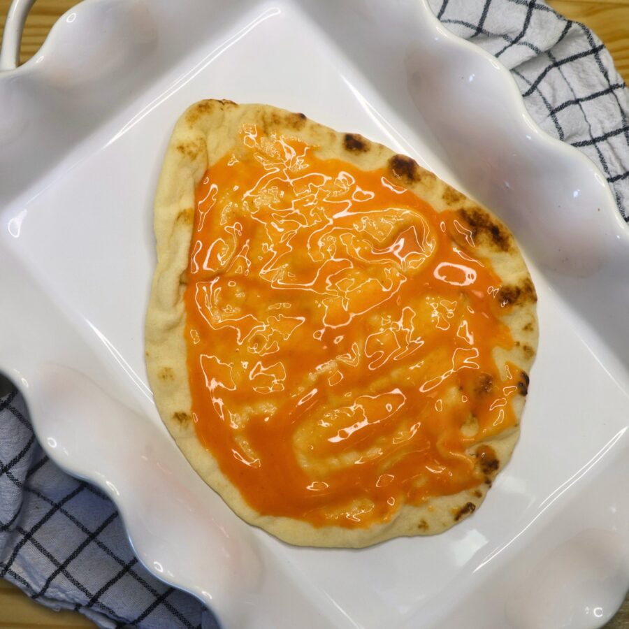 A large flatbread covered in buffalo sauce in a white baking dish