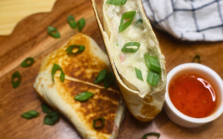 Overhead image of a crab rangoon burrito cut in half on a wooden serving tray with dipping sauce