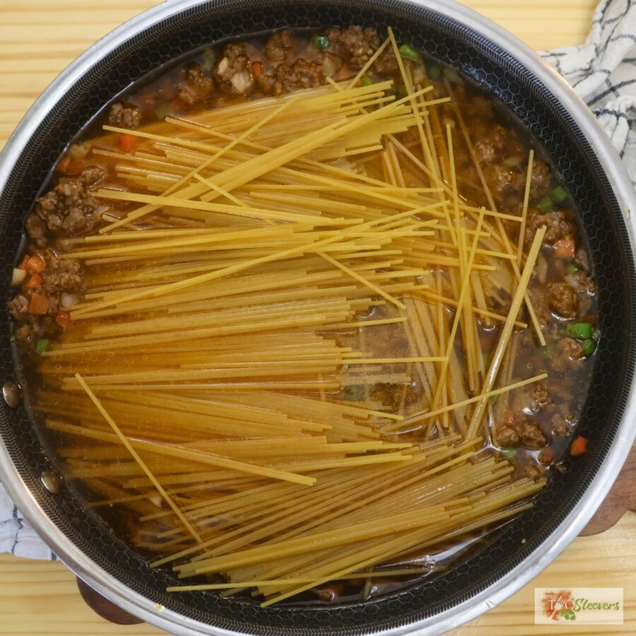 Ground beef and veggies in a skillet with broth and uncooked spaghetti noodles