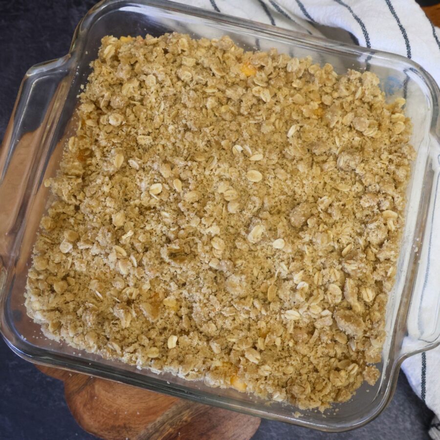 Peach crumble topping layered over peach filling and crust in a glass baking dish