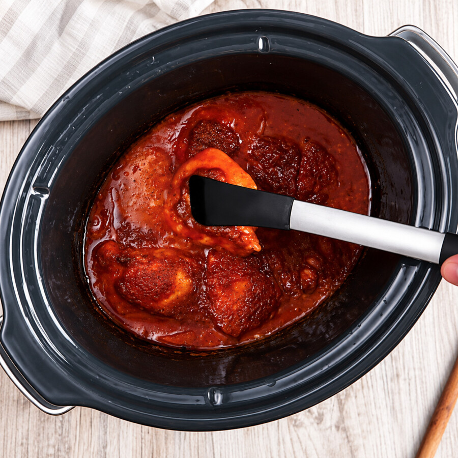 Cooked chicken thighs being removed from a slow cooker with sauce with a pair of tongs