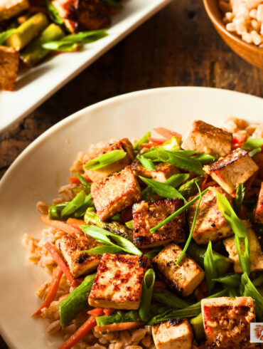 Tofu stir fry on a plate over rice with a serving dish of it next to the plate