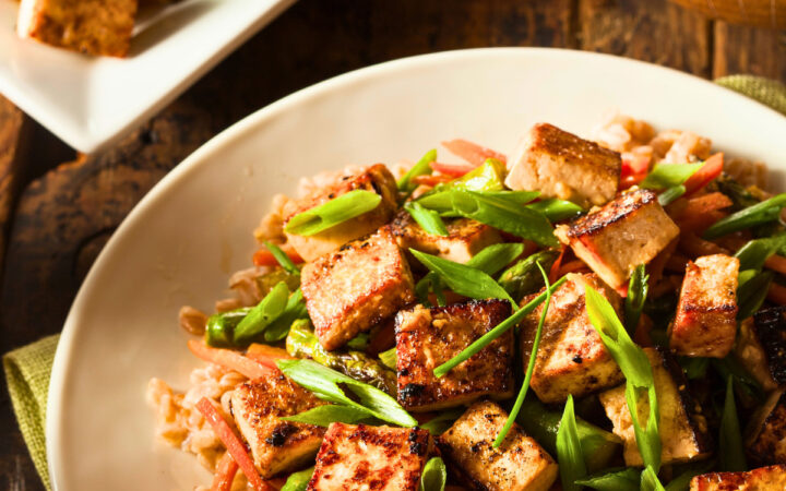 Tofu stir fry on a plate over rice with a serving dish of it next to the plate