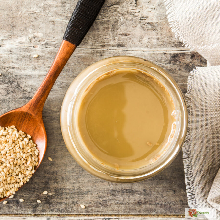 Overhead image of tahini dressing in a glass jar with a spoon of sesame seeds